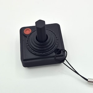 May include: A black miniature joystick with a red button and a black cord. The joystick has a hexagonal handle and a circular base with concentric rings. A retro gaming accessory.