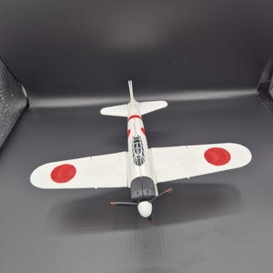 May include: A white model airplane with red accents and a red circle on each wing. The propeller is black, and the cockpit is visible. The plane is a replica of a vintage aircraft.
