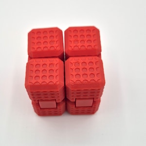May include: A red fidget cube with a textured surface and a series of connected blocks. Each block has a pattern of small, recessed circles. The cube is designed for tactile manipulation and stress relief.