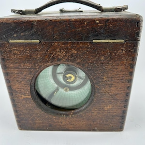 May include: A weathered wooden box with a circular window displaying a detailed instrument. The box features a leather handle and metal hinges. The instrument inside has a green and white patterned dial with a gold centre.