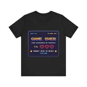 May include: A black short-sleeve t-shirt featuring a pixelated video game screen graphic. The text reads GAME OVER, TIME EXCEEDED IN TRANSIT, TTL with three red hearts, and INSERT COIN TO RETRY with two yellow coin icons and TTL=64.