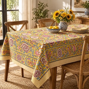 May include: A rectangular tablecloth featuring a vibrant yellow background with a repeating floral pattern in shades of red, pink, and blue. The fabric has a decorative border with a striped edge and is draped over a wooden dining table.
