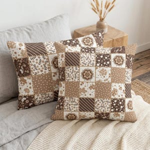 May include: Two square throw pillows featuring a patchwork design in shades of brown, beige, and white. The pattern consists of alternating squares with floral motifs, polka dots, and diagonal stripes, creating a cohesive grid-like appearance.