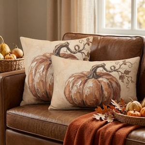 May include: Two rectangular throw pillows featuring a painted illustration of a large orange pumpkin with a dark brown stem and curling vines against a cream-colored background. The artwork displays visible brushstrokes and textured paint details.