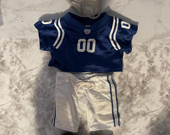 Abbigliamento Build-a-Bear - uniforme da calcio per ragazzi. Maglia blu con numeri bianchi, pantaloni da calcio bianchi, scarpe da calcio nere con tacchetti e casco in tessuto.