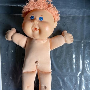 May include: A doll with a soft, light-toned fabric body and a molded vinyl head. The doll features bright blue eyes, a small open mouth, and peach-colored yarn hair styled in a top knot. The doll has a navel indentation and defined toes.