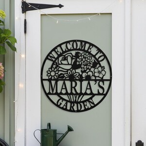 May include: Black metal garden sign with the text "WELCOME TO MARIA'S GARDEN" surrounded by floral designs. A green watering can sits on a wooden shelf below the sign. The sign is mounted on a light green door.