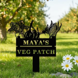 Op de afbeelding: Zwart metalen tuinbord met de tekst "MAYA'S VEG PATCH" en een decoratieve bovenkant met silhouetten van groenten. Het bord staat in een grasveld met bomen op de achtergrond en witte madeliefjes op de voorgrond.