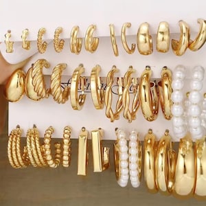 May include: A collection of gold-colored earrings in various styles, including hoops, studs, and textured designs. Some earrings feature a pearl-like white bead design. The earrings are displayed on a white card.