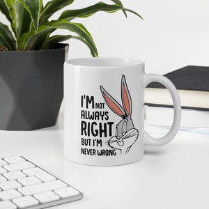 Peut inclure: Un mug en céramique blanche orné d'une illustration du personnage de dessin animé Bugs Bunny avec des oreilles aux intérieurs roses et des moustaches noires. Le texte imprimé en noir indique : I'M NOT ALWAYS RIGHT BUT I'M NEVER WRONG.
