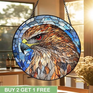 Puede incluir: Arte de ventana de vidriera redonda que presenta la cabeza detallada de un halcón con ojos rojos, pico amarillo y plumas marrones y blancas. El fondo es un mosaico de vidrio azul y blanco. El texto "BUY 2 GET 1 FREE" está en la parte inferior.