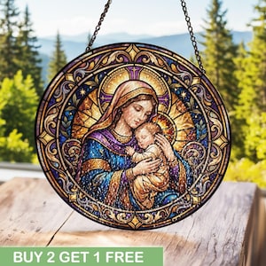 May include: A circular stained glass suncatcher featuring a depiction of a person holding a baby. The artwork uses deep blue, purple, and gold tones with intricate decorative borders and a golden halo effect behind the figures, suspended by a chain.
