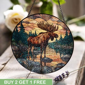 Könnte beinhalten: Runde Glasmalerei mit einem Elch, der in einem See steht, umgeben von Bäumen und einem Sonnenuntergang. Das Kunstwerk hat einen schwarzen Metallrahmen und eine Aufhängekette. Der Text "BUY 2 GET 1 FREE" befindet sich unten.