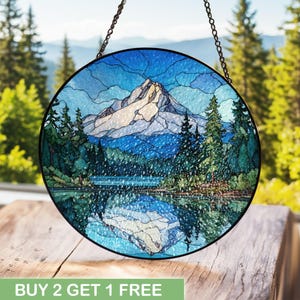 Könnte beinhalten: Rundes Buntglasfenster mit einer Berglandschaft mit See und Bäumen. Das Kunstwerk zeigt Blau-, Grün- und Weißtöne, mit einem schwarzen Metallrahmen und einer Kette. Der Text "BUY 2 GET 1 FREE" befindet sich unten.