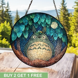 以下が含まれることがあります： 丸いステンドグラスパネル。木の下にいる漫画のキャラクターと満月が描かれています。キャラクターはグレーと白で、大きな目があります。葉は青と緑の様々な色合いです。