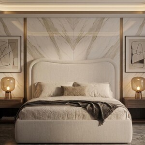 May include: A beige upholstered bed with a curved headboard, two dark wood nightstands, and two amber glass table lamps. The bed is dressed in neutral linens with a dark throw blanket. Two framed abstract line art prints hang on a marble wall.