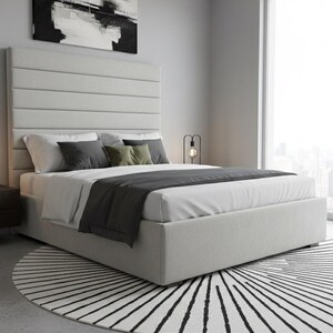 May include: A modern bedroom scene featuring a light gray upholstered bed with a tall, horizontally paneled headboard. The bed is made with white linens, dark gray and olive green pillows, and a dark gray throw. A black and white rug sits at the foot of the bed.