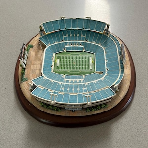 May include: A miniature scale model of the Jacksonville Jaguars football stadium featuring teal seating, a green field with white yard lines, and the text Jacksonville and Jaguars printed on the turf, all mounted on a circular wooden base.