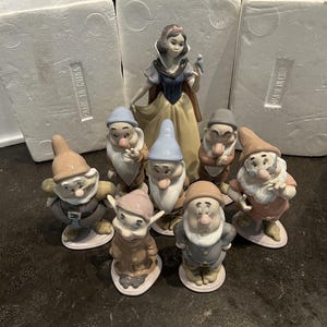Può includere: Un set di statuine in porcellana raffigurante Biancaneve in un abito blu e giallo con un piccolo uccellino, accompagnata da sette statuine di nani con abiti e cappelli di vari colori, su basi bianche con imballaggio in schiuma.