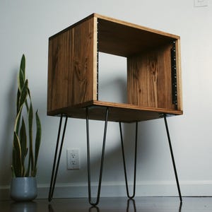 May include: A rectangular wooden storage unit with a visible grain pattern, featuring an open front and interior black metal rack rails. The unit is supported by four thin, black metal hairpin legs that extend outward from the base.