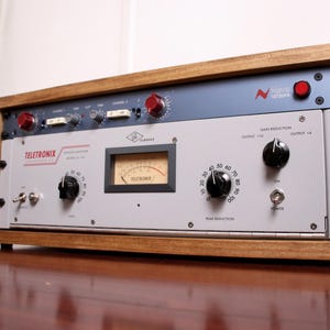 May include: A vintage Teletronix LA-2A audio compressor in a wooden frame. The device has a gray faceplate with black knobs, a VU meter, and red accents. The frame is made of light brown wood.