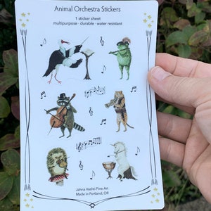 May include: A sheet of stickers featuring an animal orchestra. The stickers are colorful and feature a variety of animals playing musical instruments. The text on the sheet reads "Animal Orchestra Stickers" and "1 sticker sheet, multipurpose, durable, water resistant".