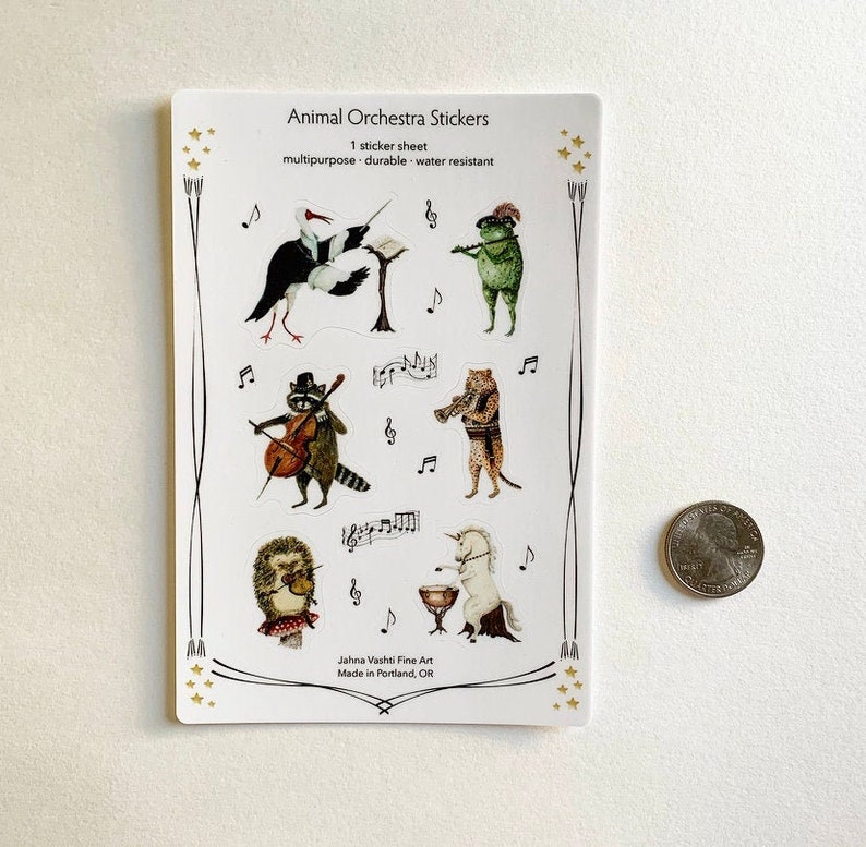 May include: A sheet of stickers featuring various animals playing musical instruments. The animals include a raccoon playing cello, a hedgehog playing violin, a unicorn playing drums, a frog playing flute, a bird playing a conductor's baton, and a cheetah playing trumpet. The stickers are labeled "Animal Orchestra Stickers" and "Jahna Vashti Fine Art, Made in Portland, OR".