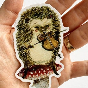 May include: A sticker featuring a cartoon hedgehog playing a violin while sitting on a red and white polka dot mushroom.