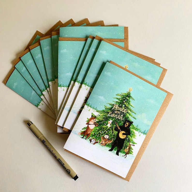 May include: A pack of 10 Christmas cards with a brown envelope. The card features a watercolour illustration of a bear playing a banjo with other woodland animals around a Christmas tree. The text on the tree reads "Peace on Earth".