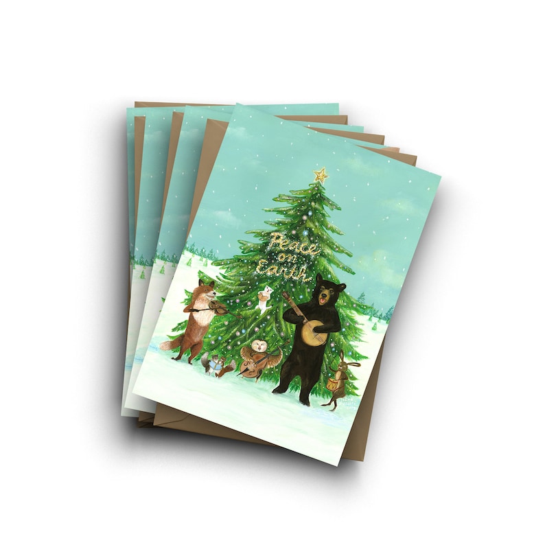 May include: A set of Christmas cards featuring a winter scene with a green pine tree decorated with baubles and a star. A black bear plays a banjo, a fox plays a violin, and other woodland creatures gather around the tree. The text "Peace on Earth" is written in gold glitter on the tree.