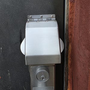 May include: A brushed metal door lock with a protective cover. The lock is mounted on a dark gray door, with a red-brown wall visible on the right side. The cover is rectangular with rounded edges and is attached with a hinge.