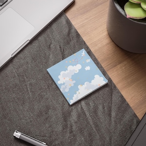 May include: A square notepad with a light blue background featuring white clouds, yellow stars, and a small airplane with a heart-shaped trail. The notepad is on a dark gray surface, near a laptop and a pen.