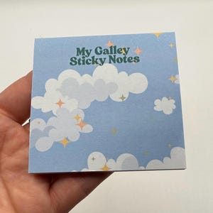 May include: Square sticky notes with a light blue background featuring white clouds and sparkling stars. The text "My Galley Sticky Notes" is printed in green and white. The notes are held in a hand.