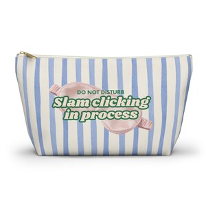 May include: A rectangular cosmetic bag with a gold zipper, featuring a blue and white striped pattern. The bag has a pink eye mask graphic with the text "DO NOT DISTURB Slam clicking in process" in green.