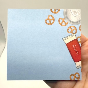 May include: A light blue square notepad with a whimsical design. The notepad features illustrations of pretzels, a can, and a red biscotti package. The word "Biscotti" is printed on the package. The notepad is held by a hand.