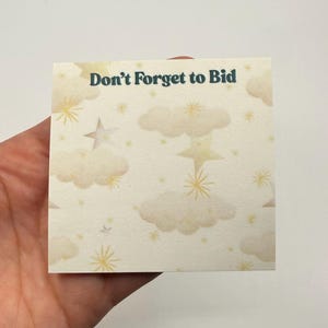 May include: A square sticky note with a cream background featuring a pattern of yellow stars and clouds. The text "Don't Forget to Bid" is printed in dark teal at the top. The note is held in a hand.