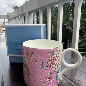 May include: A pink ceramic mug with a floral design and gold trim. The mug features a white handle and is next to a blue box with the text "WEDGWOOD" and "LILAC CRANE". The mug is on a wooden surface.
