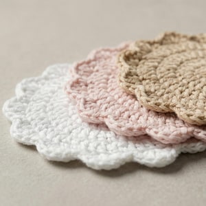 Op de afbeelding: Drie gehaakte onderzetters in wit, roze en beige. Elke onderzetter heeft een geschulpte rand en een spiraalpatroon. De onderzetters zijn gemaakt van dik, getextureerd garen.