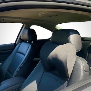 Pode incluir: Encosto de cabe&ccedil;a preto para banco de carro com uma almofada de apoio para o pesco&ccedil;o. O encosto de cabe&ccedil;a est&aacute; preso a um banco de carro de couro preto. O interior do carro &eacute; vis&iacute;vel, com um fundo branco vis&iacute;vel atrav&eacute;s das janelas.