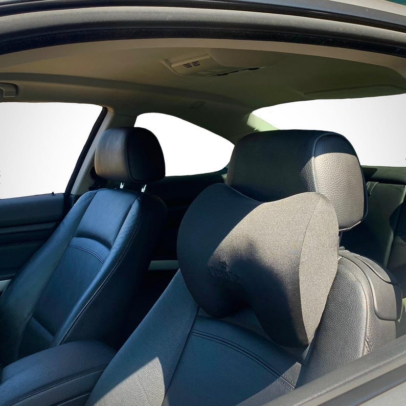 Pode incluir: Encosto de cabe&ccedil;a preto para banco de carro com uma almofada de apoio para o pesco&ccedil;o. O encosto de cabe&ccedil;a est&aacute; preso a um banco de carro de couro preto. O interior do carro &eacute; vis&iacute;vel, com um fundo branco vis&iacute;vel atrav&eacute;s das janelas.