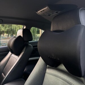Pode incluir: Almofada preta para encosto de cabe&ccedil;a de carro com design curvo. A almofada &eacute; presa a um assento de carro de couro preto. O encosto de cabe&ccedil;a tem a marca "CuteCat" impressa.