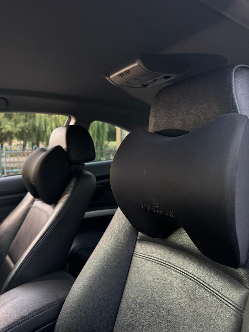 Pode incluir: Almofada preta para encosto de cabe&ccedil;a de carro com design curvo. A almofada &eacute; presa a um assento de carro de couro preto. O encosto de cabe&ccedil;a tem a marca "CuteCat" impressa.