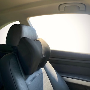 Pode incluir: Almofada preta para encosto de cabe&ccedil;a de carro com um design contornado, proporcionando suporte para o pesco&ccedil;o. A almofada &eacute; presa a um assento de carro de couro preto. O encosto de cabe&ccedil;a est&aacute; em frente a uma janela brilhante.