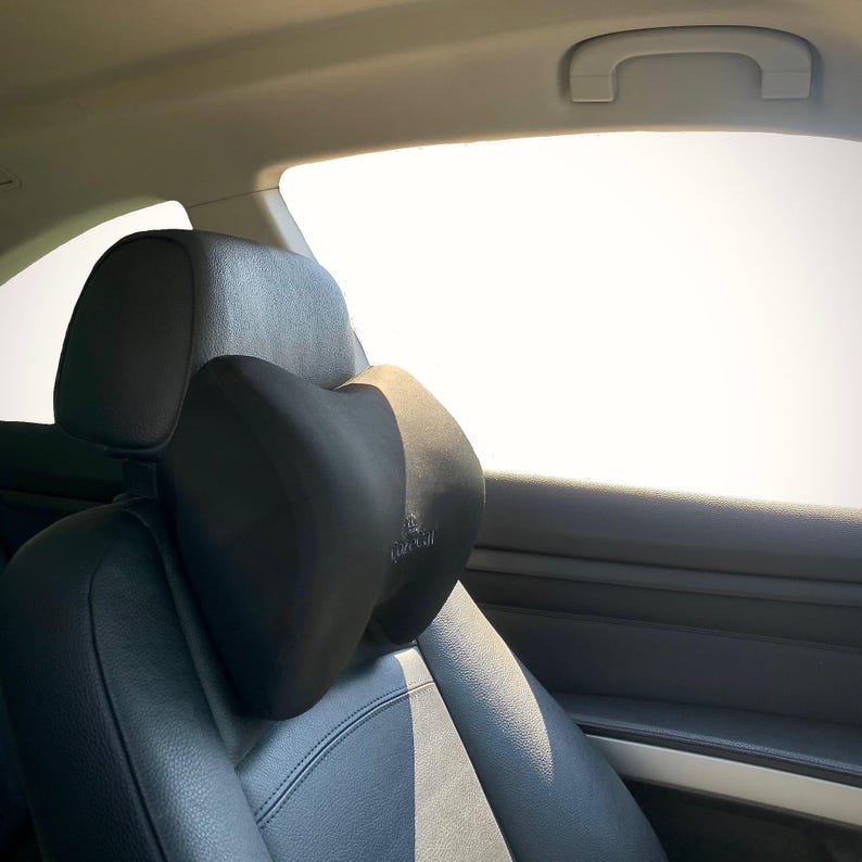 Pode incluir: Almofada preta para encosto de cabe&ccedil;a de carro com um design contornado, proporcionando suporte para o pesco&ccedil;o. A almofada &eacute; presa a um assento de carro de couro preto. O encosto de cabe&ccedil;a est&aacute; em frente a uma janela brilhante.