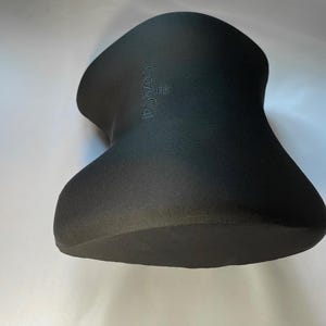 Pode incluir: Uma almofada de pesco&ccedil;o preta com uma forma incomum de ampulheta. A almofada tem uma superf&iacute;cie texturizada e a palavra "CozyCal" est&aacute; em relevo no topo. Projetada para suporte e conforto do pesco&ccedil;o.