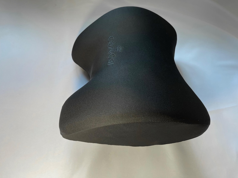 Pode incluir: Uma almofada de pesco&ccedil;o preta com uma forma incomum de ampulheta. A almofada tem uma superf&iacute;cie texturizada e a palavra "CozyCal" est&aacute; em relevo no topo. Projetada para suporte e conforto do pesco&ccedil;o.
