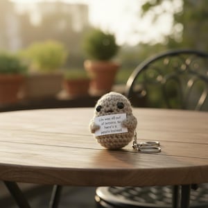 May include: A crocheted potato keychain in shades of tan and brown, holding a small sign that reads "Life was all out of lemons, so here's a potato instead." The keychain is on a wooden table, with a blurred outdoor background.
