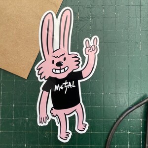 May include: A pink cartoon rabbit sticker with a black t-shirt that says "METAL". The rabbit is giving a rock and roll hand gesture. The sticker has a white border and is on a green surface.
