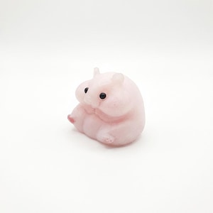 May include: A small, pink hamster figurine with black eyes and tiny pink feet. The hamster is in a curled-up position, appearing to be resting or sleeping. The figurine is made of a smooth material.