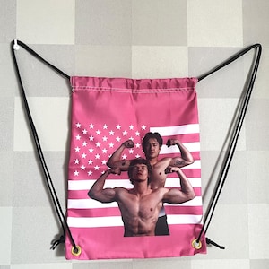 May include: Pink drawstring bag featuring a graphic of two muscular men flexing their biceps. The bag has a pink American flag design with white stars and stripes. Black drawstrings and gold eyelets complete the design.